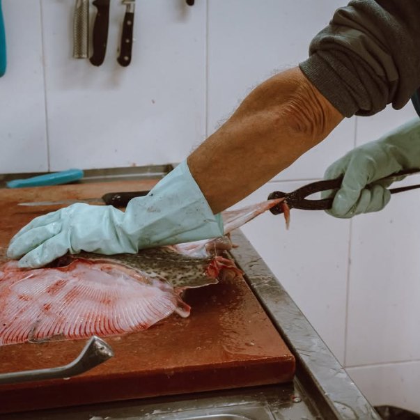 Manos con guantes limpian un pescado plano sobre una tabla.